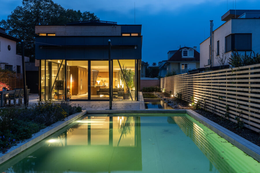 Die beleuchtete Gartenansicht eines modernen Wohnhauses zur blauen Stunde mit einem hell erstrahlten Innenraum und einem beleuchteten Naturpool im Vordergrund.
