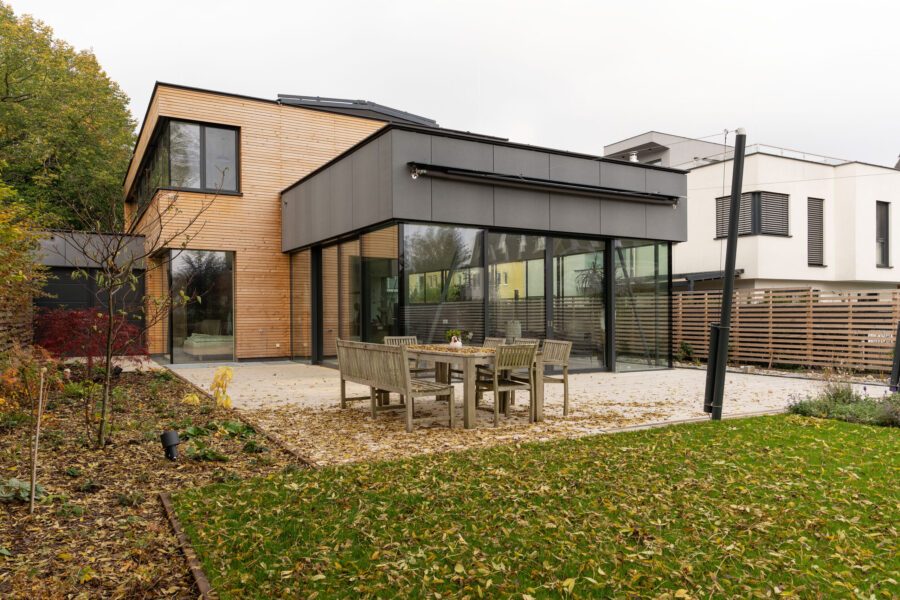 Die Seitenansicht eines modernen Wohnhauses im Herbst mit einer gepflasterten Terrasse, einer Holzsitzgruppe, großen Glasfronten und einer Fassade aus Holz und anthrazitfarbenen Platten.