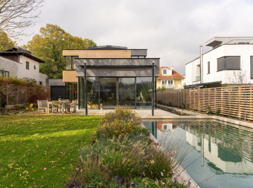 Die Gartenansicht eines modernen, mehrstöckigen Wohnhauses mit einer großen Glasfront, einer gepflasterten Terrasse mit Sitzgruppe, einer Rasenfläche und einem Naturpool im Vordergrund.