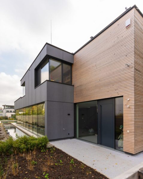 Die Außenansicht eines modernen, mehrstöckigen Wohnhauses mit einer Fassadenkombination aus hellen Holzlatten und dunkelgrauen Platten, großen Fensterfronten und einem angrenzenden Wasserbecken.