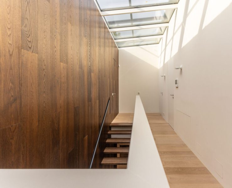Ein moderner, lichtdurchfluteter Flur mit einem Glasdach, einer dunkelbraunen Holzwand auf der linken Seite und einer Treppe, die nach unten führt.