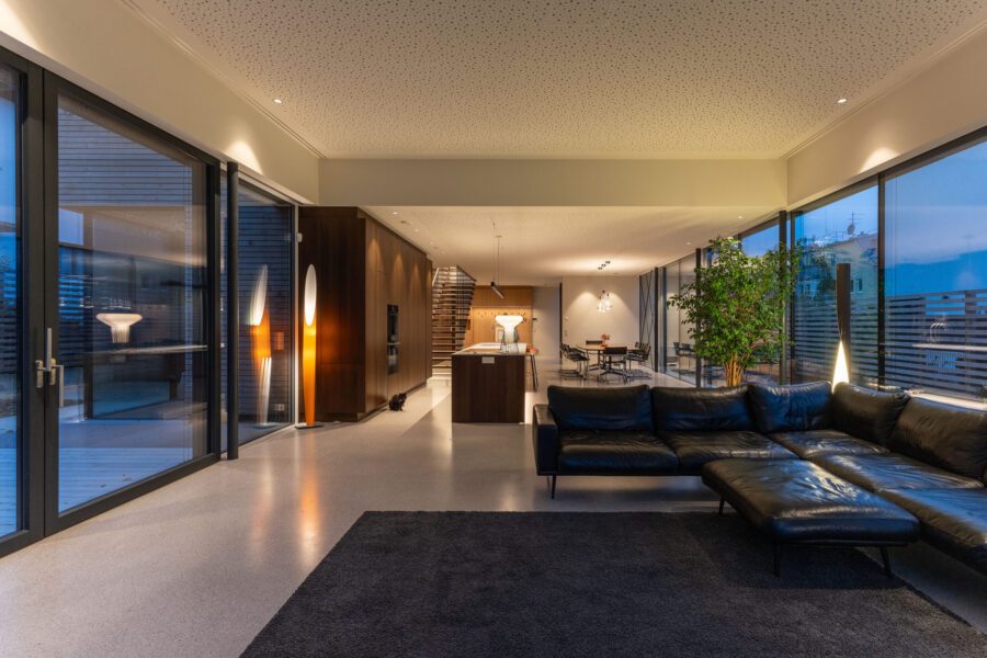 Helles, modernes Wohnzimmer mit großen Fensterfronten links und rechts. Im Vordergrund liegt ein Teppich, auf dem eine Leder-Sitzgarnitur steht. Im Hintergrund führt eine Treppe nach oben, daneben befindet sich ein Essbereich mit Sitzgelegenheiten.