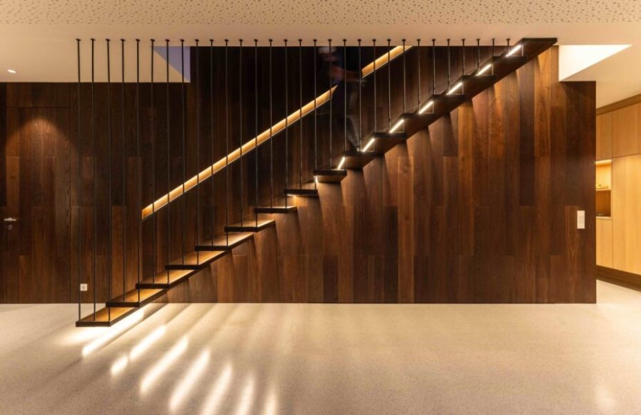 Moderne, schwebende Holztreppe mit integrierter LED-Stufenbeleuchtung vor einer dunklen Holzwand.